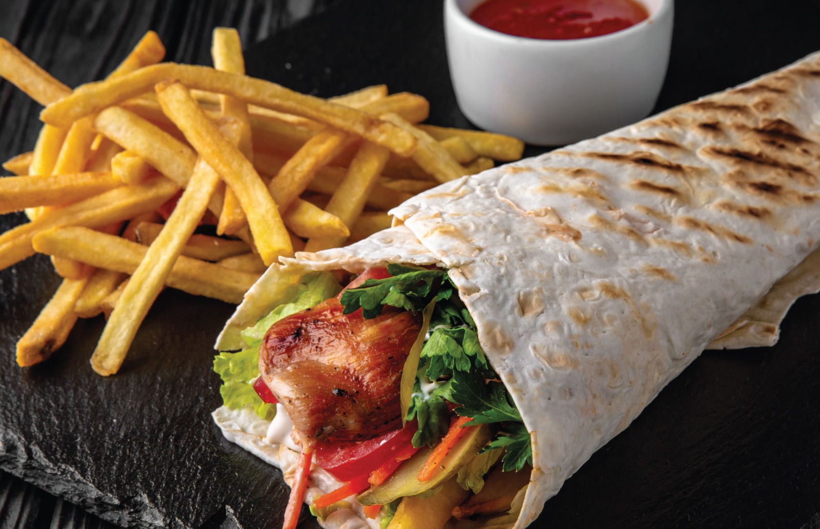 Slate with a grilled chicken wrap filled with salad and mayo, portion of fries on the side with a tomato sauce for dipping
